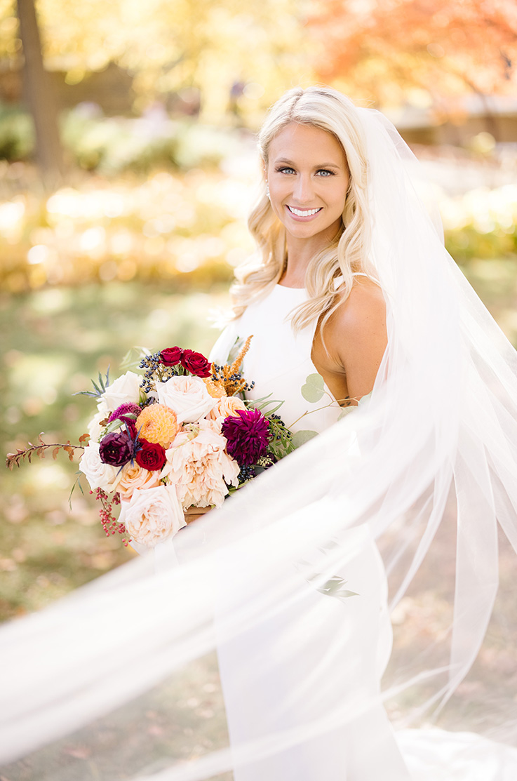 A bride in a sleeveless wedding dress holds a veil and smiles, while her veil flows out in front of her as she stands outside.