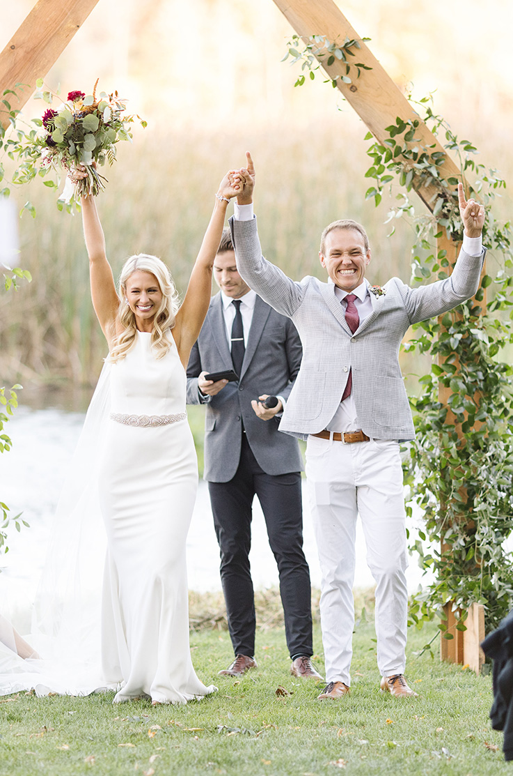 A bride in a sleeveless wedding dress and a veil, and a groom in a suit, raise their hands together and smile after saying their vows outside in front of a lake, a man, and a ceremony decoration.
