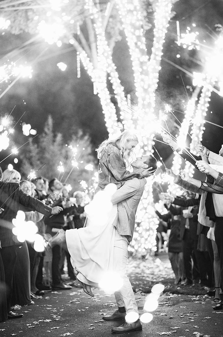 A groom in a suit lifts a bride in a wedding dress and jacket while fireworks go off in the background and wedding guests hold up sparklers.