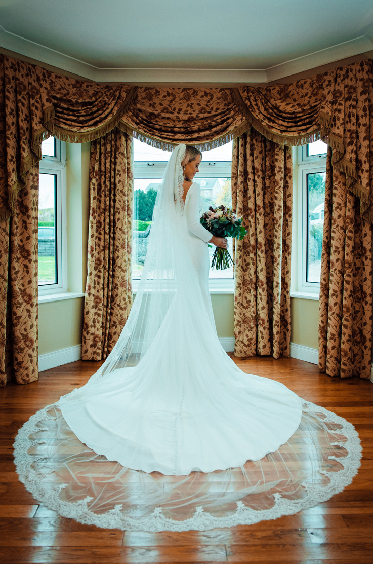 A bride in a long sleeve wedding dress holds a bouquet and poses indoors, in front of a windowed alcove, with her train and veil fanned out behind her.