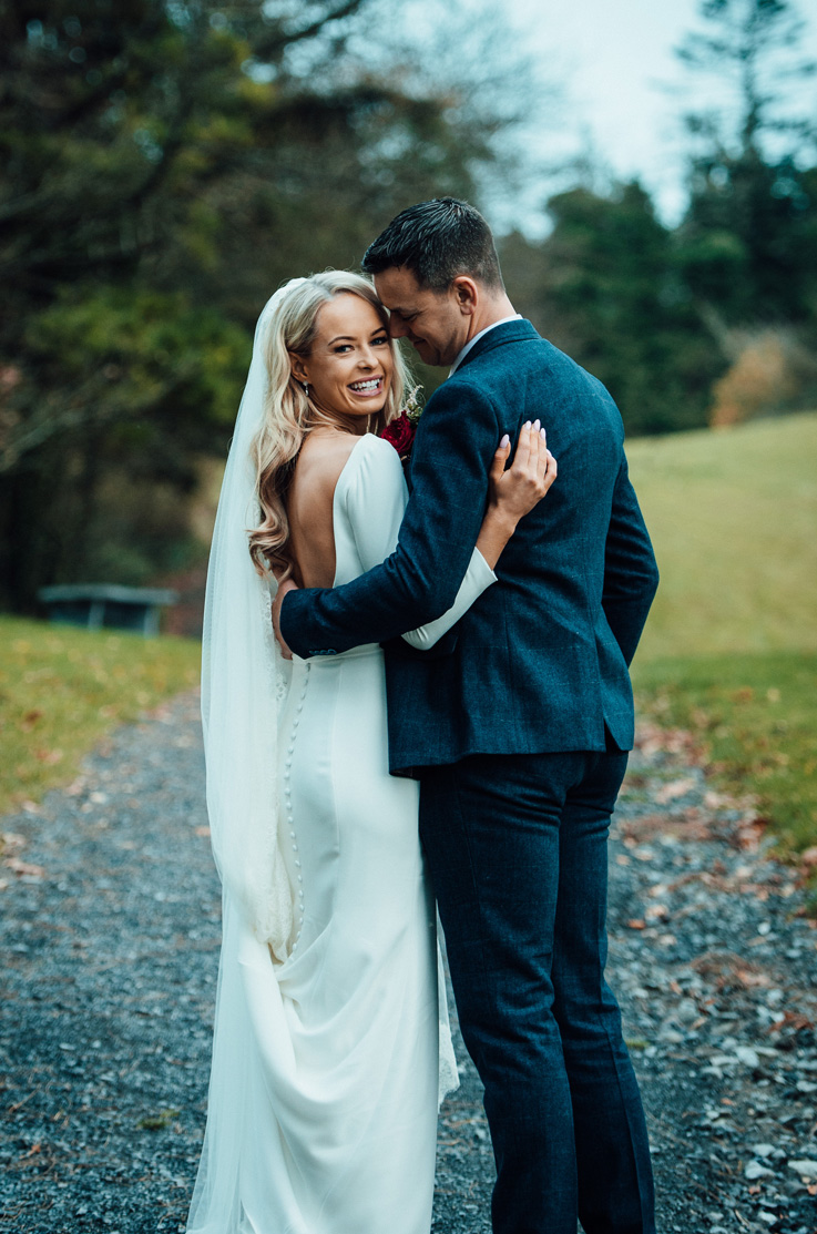 A bride in a long sleeve wedding dress looks over her shoulder and smiles while a groom in a suit holds her close, both of them standing on a gravel path outside with trees in the background.