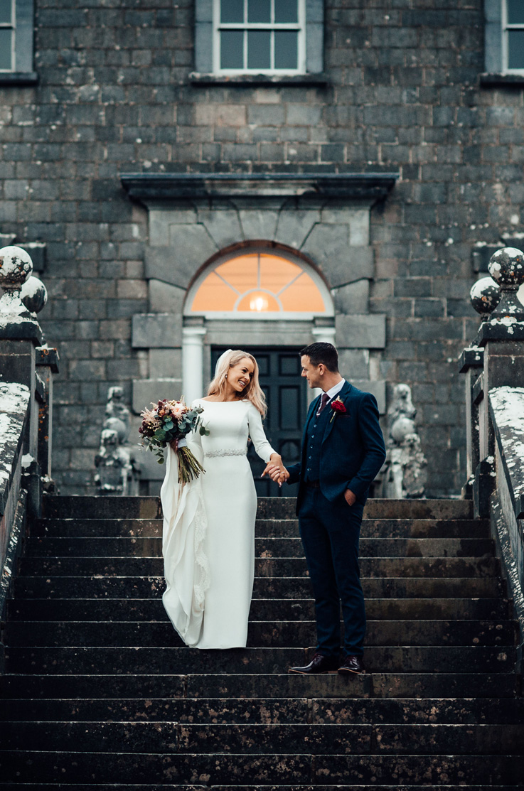 A bride in a long sleeve wedding dress holds a bouquet in one hand, and the hand of a groom in a suit in the other, while they smile at each other and stand on a staircase in front of a stone building.