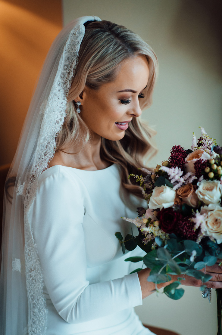 A bride in a long sleeve wedding dress and veil holds a bouquet and looks down at it while standing indoors.
