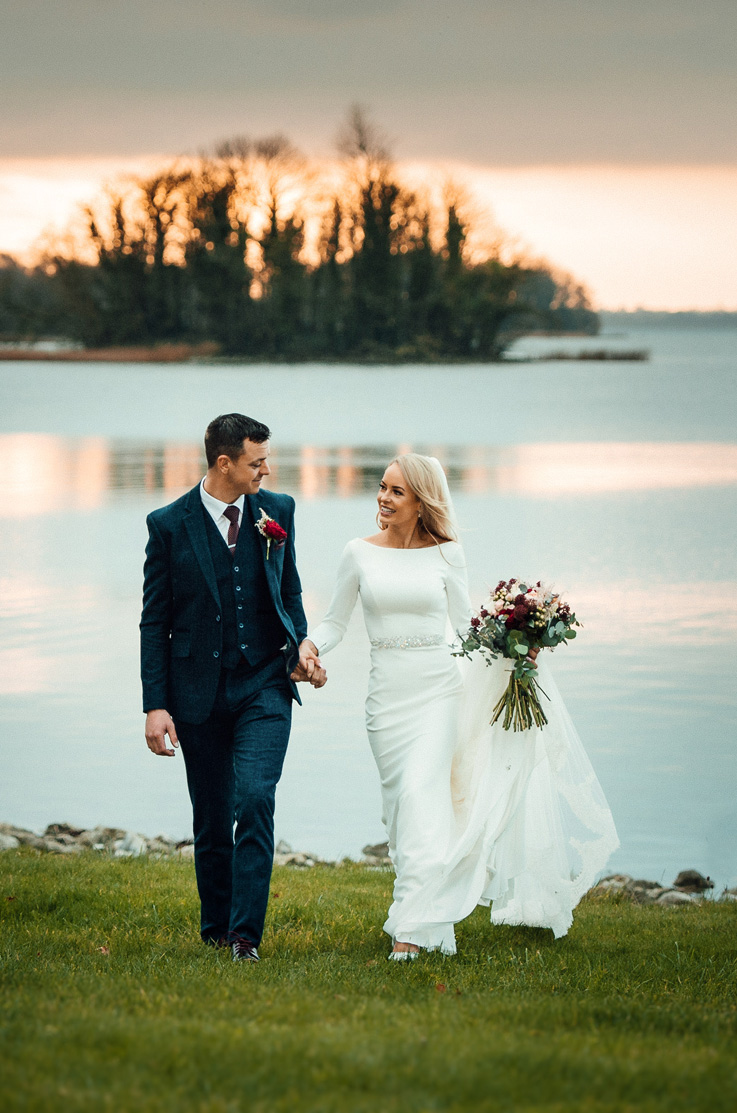 A bride in a long sleeve wedding dress holding a bouquet, and a groom in a suit, stand in front of a lake at sunset with a small island in the distance. 