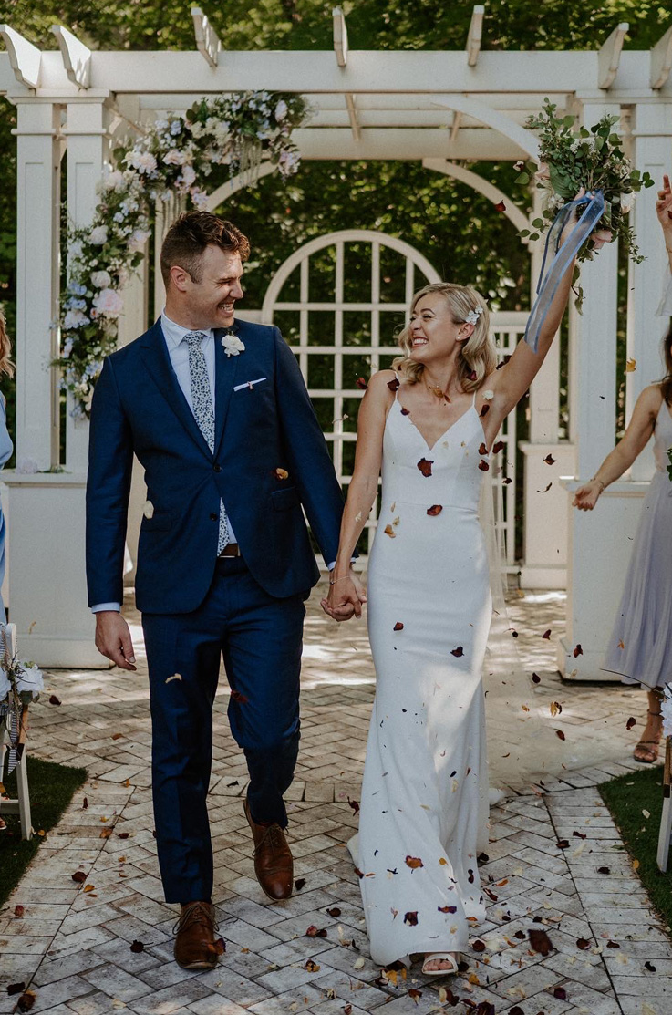 A bride in a V-neckline wedding dress and a groom in a suit walk down the aisle and smile at each other while wedding guests throw flower petals at the newlywed couple.