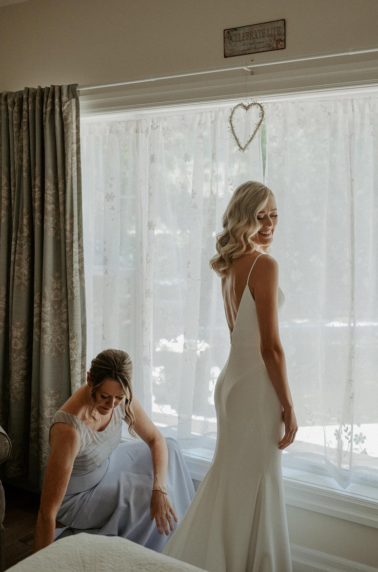 A bride in a fitted wedding dress stands by a window with sheer curtains, as she turns her head and smiles, while a woman in a dress crouches on the ground beside her.