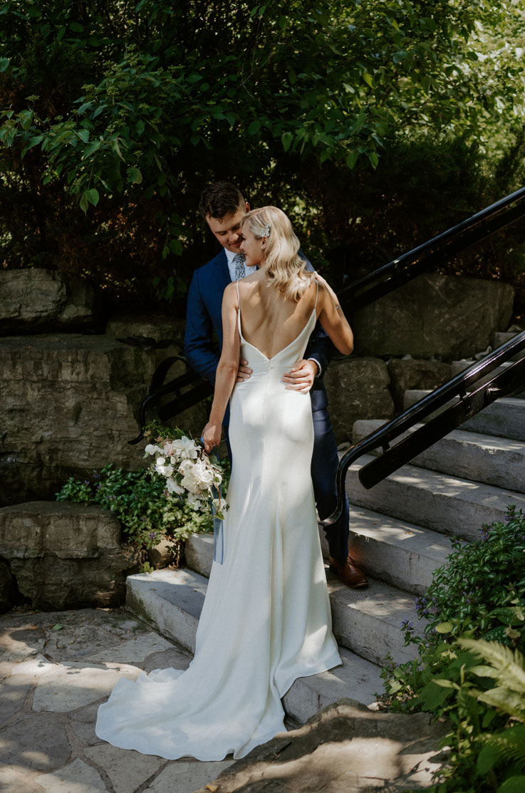 A bride in a fitted wedding dress and a groom in a suit hold each other and stand at the bottom of an outdoor staircase, with a rock wall and trees behind them.
