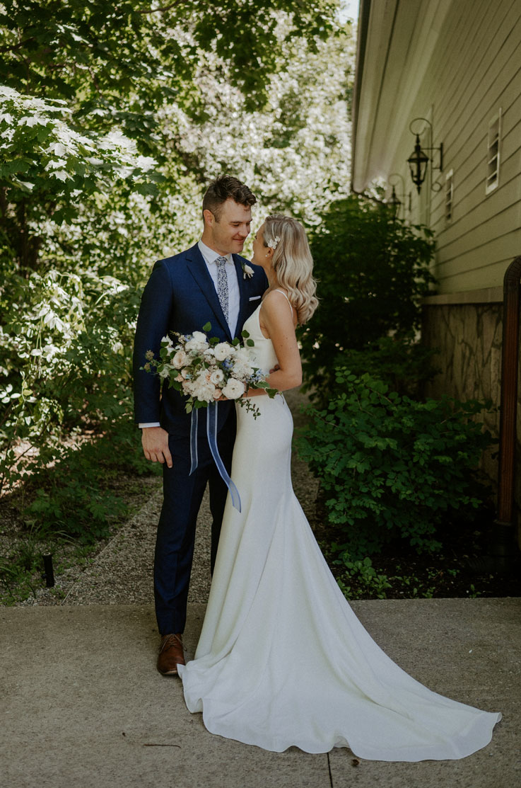 A bride in a fitted wedding dress and a groom in a suit stand off to the side of a house on a path, while looking at each other and smiling.