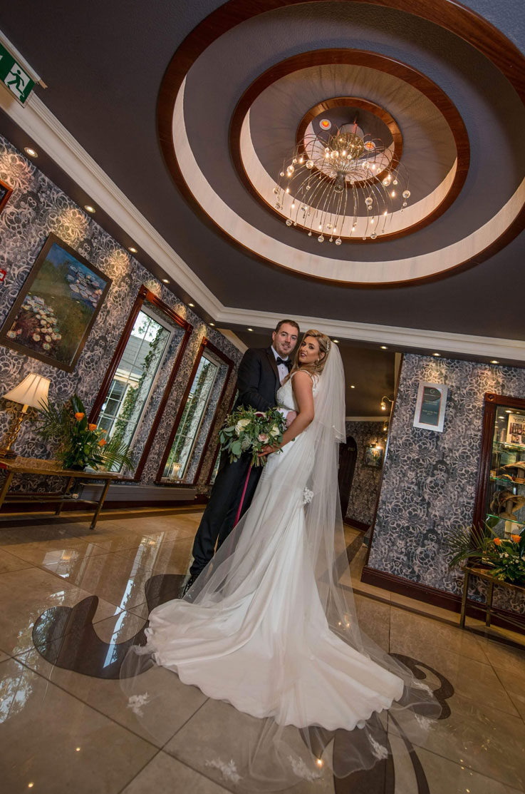 A bride in a sleeveless wedding dress holding a bouquet and a groom in a suit hold each other and smile while standing indoors under a chandelier, with the bride's dress train and veil fanned out in front of her.
