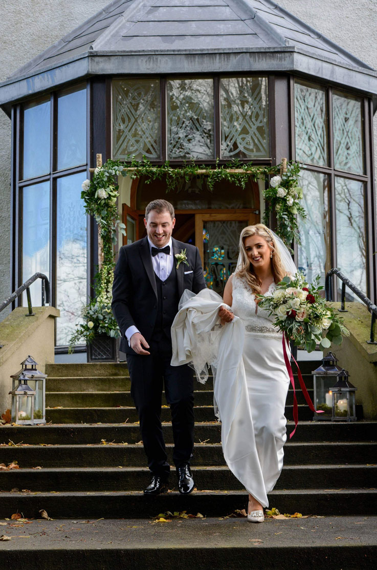 A bride in a sleeveless wedding dress and veil, holding a bouquet, and a groom stand at the bottom of a staircase in front of a building with a small sunroom entrance.
