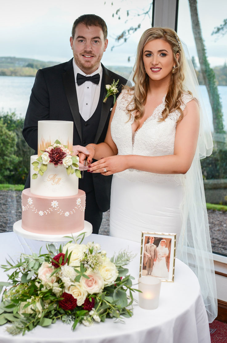 A bride in a sleeveless wedding dress and a groom in a suit stand in front of large windows, behind a small table with a tiered wedding cake on it. They smile and prepare to cut the cake.
