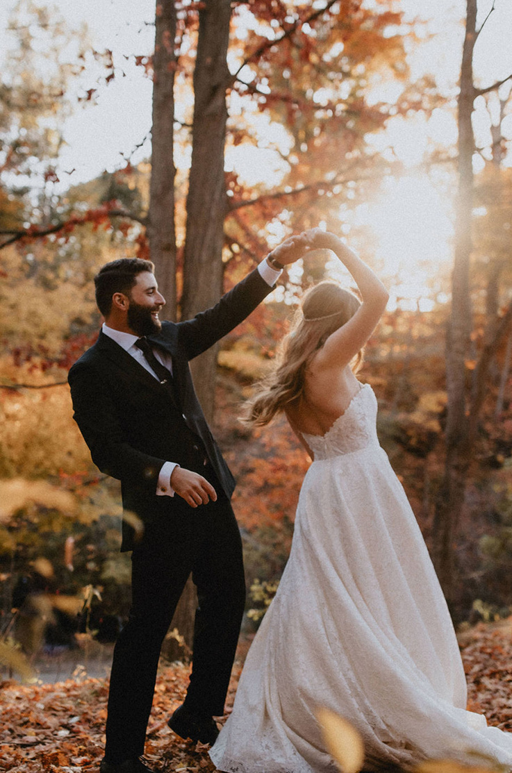 A groom in a suit spins a bride in a lace wedding dress while in a forest, with the sun behind them, visible through the trees.