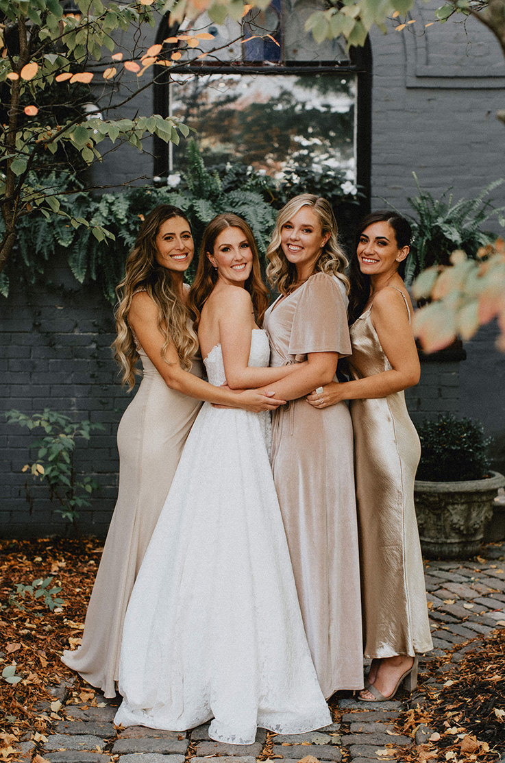 A bride in a lace wedding dress and three bridesmaids huddle together and smile while standing on a stone path outside of a brick building.