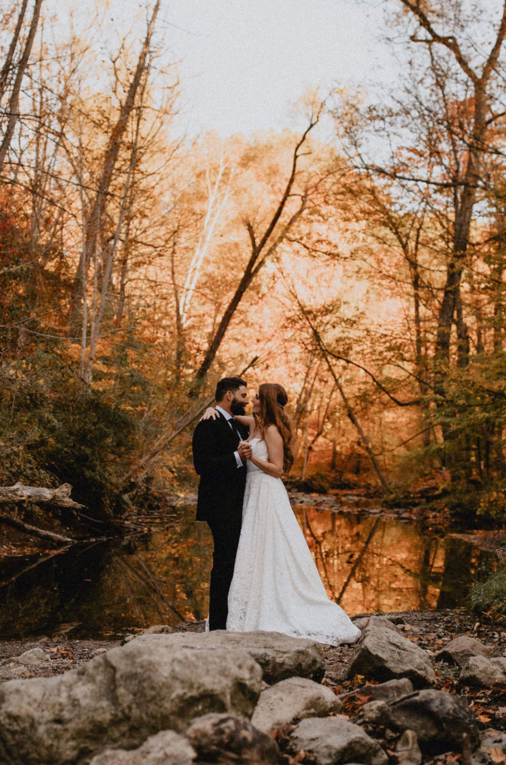 A bride in a lace wedding dress and a groom in a suit hold each other and lean in for a kiss, as they stand in a forest in front of a small pond.