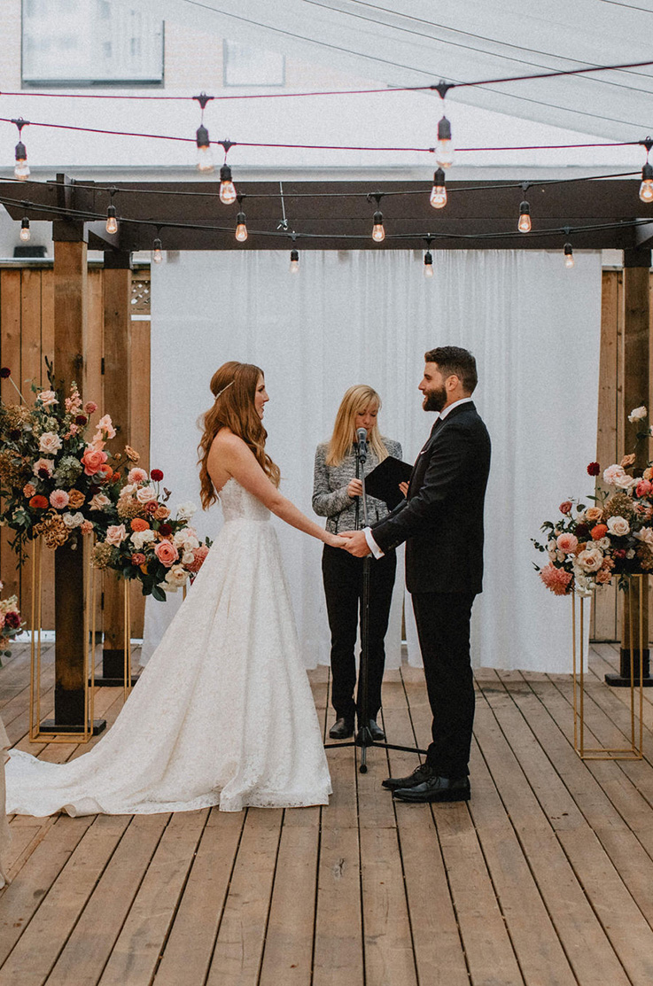 A bride in a lace wedding dress and a groom in a suit hold hands while facing each other in front of a woman talking into a microphone and a curtain backdrop, while standing outside on a wooden patio.