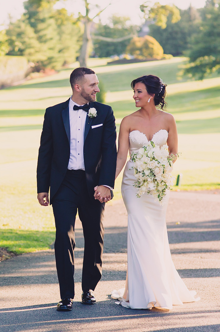 A bride in a strapless sweetheart wedding dress holds a bouquet and walks hand in hand down a path with a groom in a suit, while they look at each other and smile.