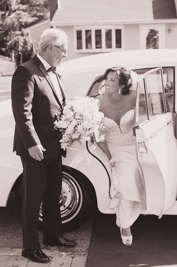 A bride in a strapless sweetheart wedding dress smiles and holds a bouquet while getting out of a vintage car, a man in a suit waits to greet her.