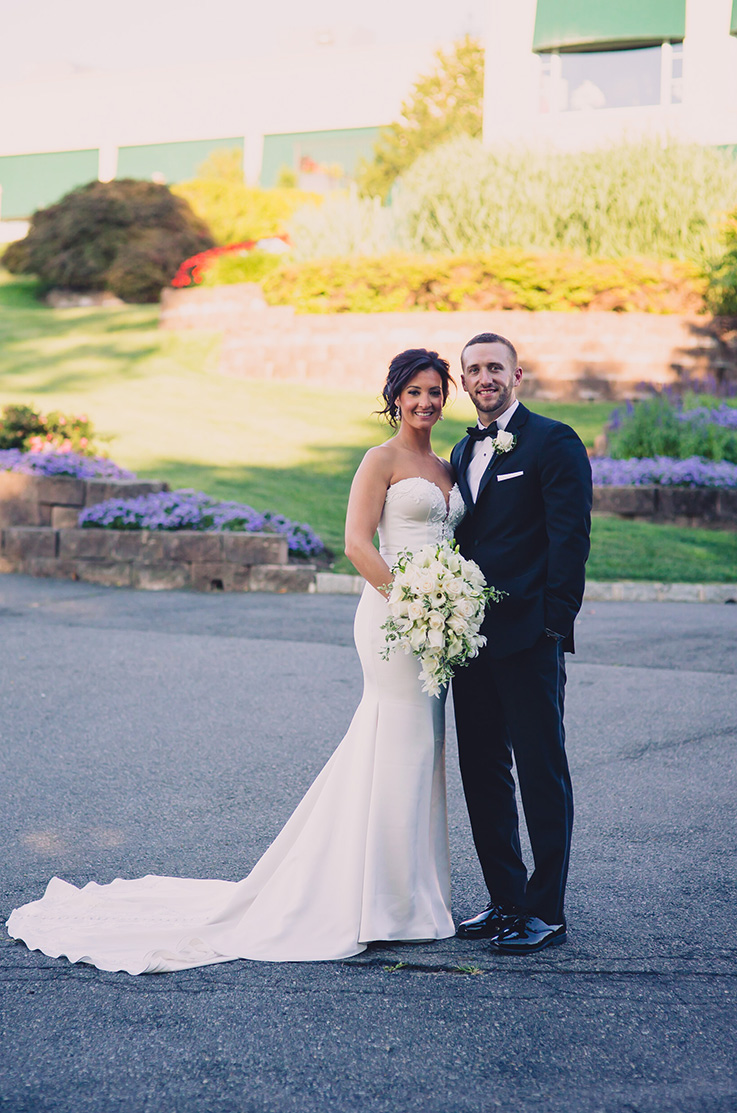 A bride in a strapless sweetheart wedding dress and a groom in a suit smile and pose in a paved area in front of a garden and a building.