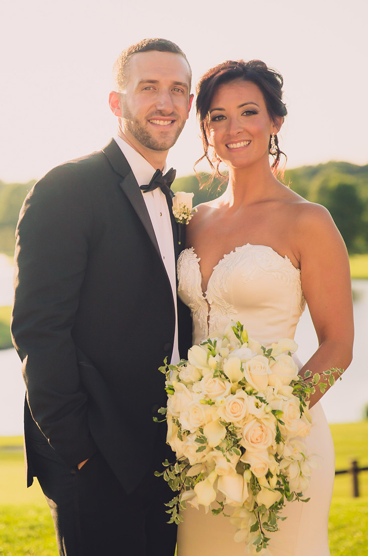 A bride in a strapless sweetheart wedding dress holds a bouquet while standing outside in front of a pond with a groom in a suit, both of them smiling.
