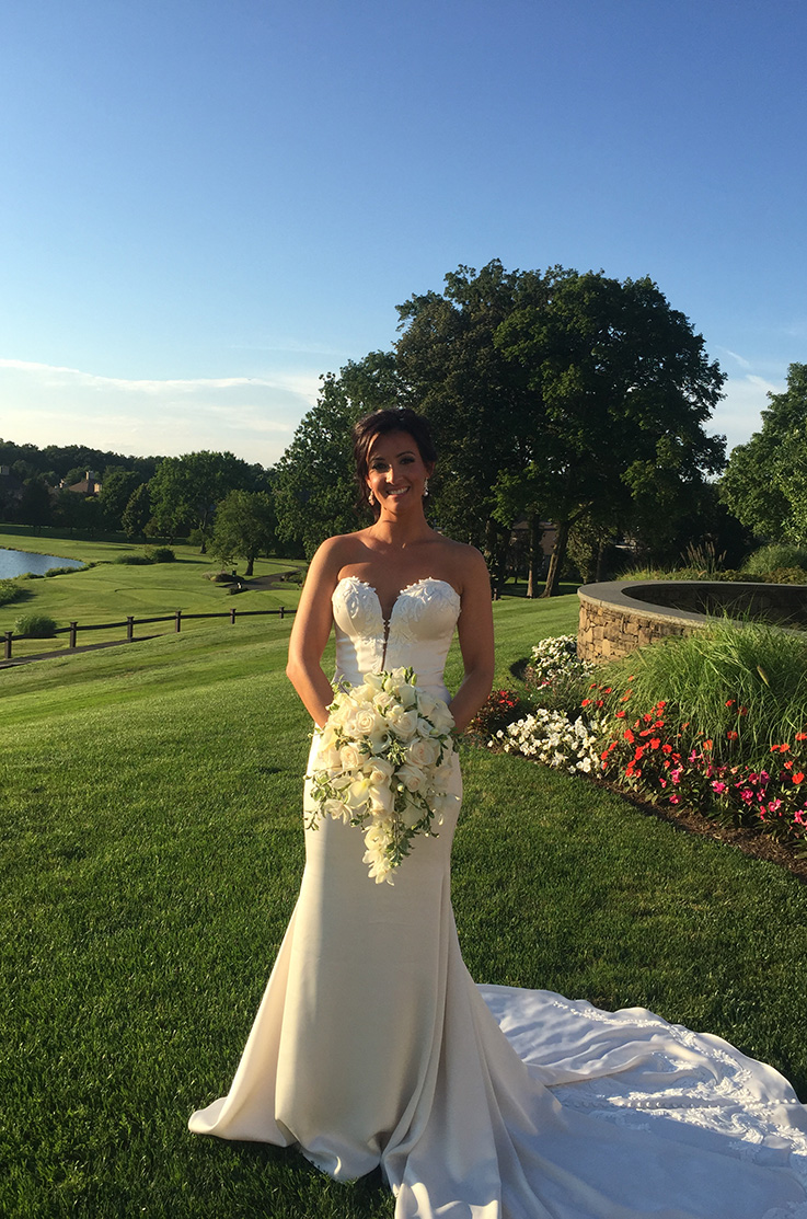 A bride in a strapless sweetheart wedding dress smiles and holds a bouquet while standing in a grass field with flowers, a fence, and trees in the background. 