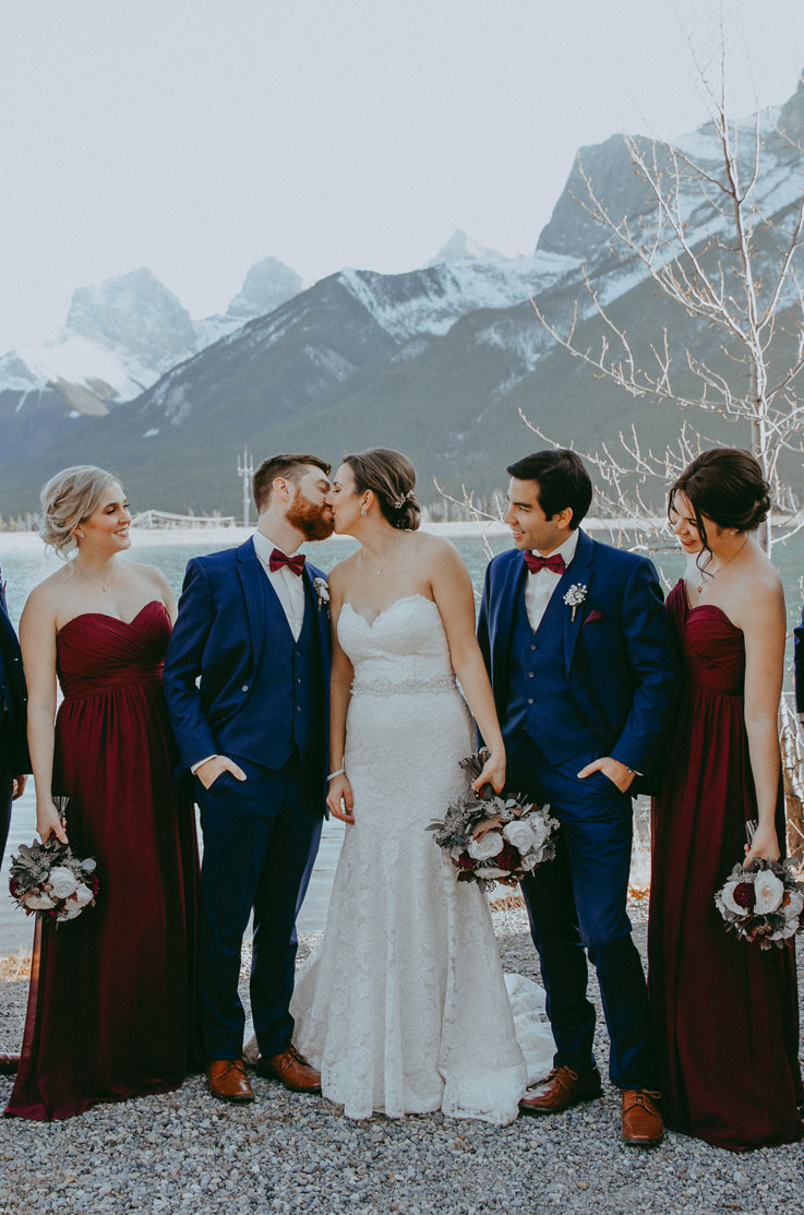 A bride in a sweetheart lace wedding dress and a groom in a suit kiss while their bridal party watches and smiles, they are all outdoors with mountains in the background.