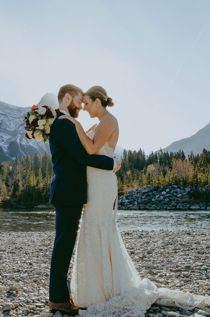 A bride in a lace wedding dress and a groom in a suit hold each other close while standing on the rocky shore of a river, with mountains and trees in the distance behind them.
