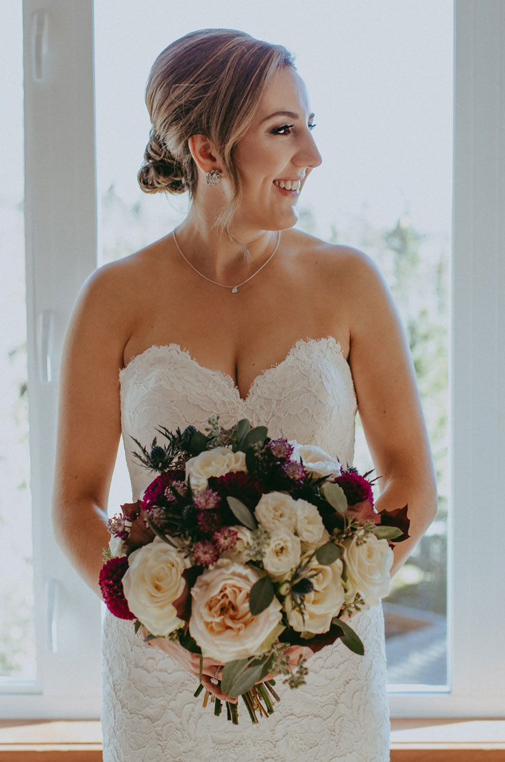 A bride in a sweetheart lace wedding dress holds a bouquet and looks to the side while smiling, as she stands indoors in front of a window.