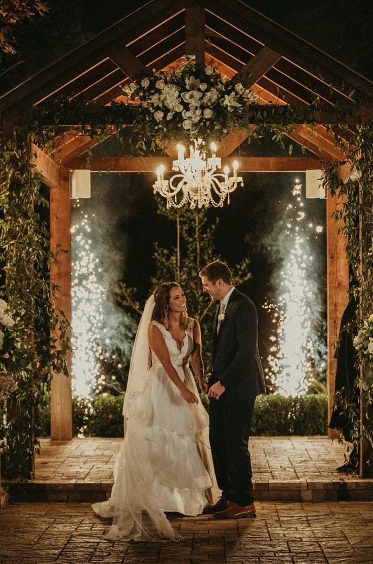 A bride in a tulle wedding dress and a groom in a suit stand under a decorated pitched pergola while looking at each other, as small pyrotechnics go off behind them.