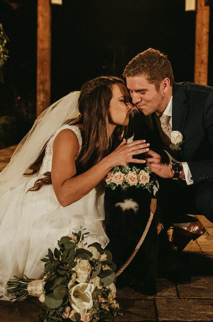 A bride in a wedding dress and a groom in a suit crouch down and give a good sweet dog kisses, while all are outside on a stone patio.