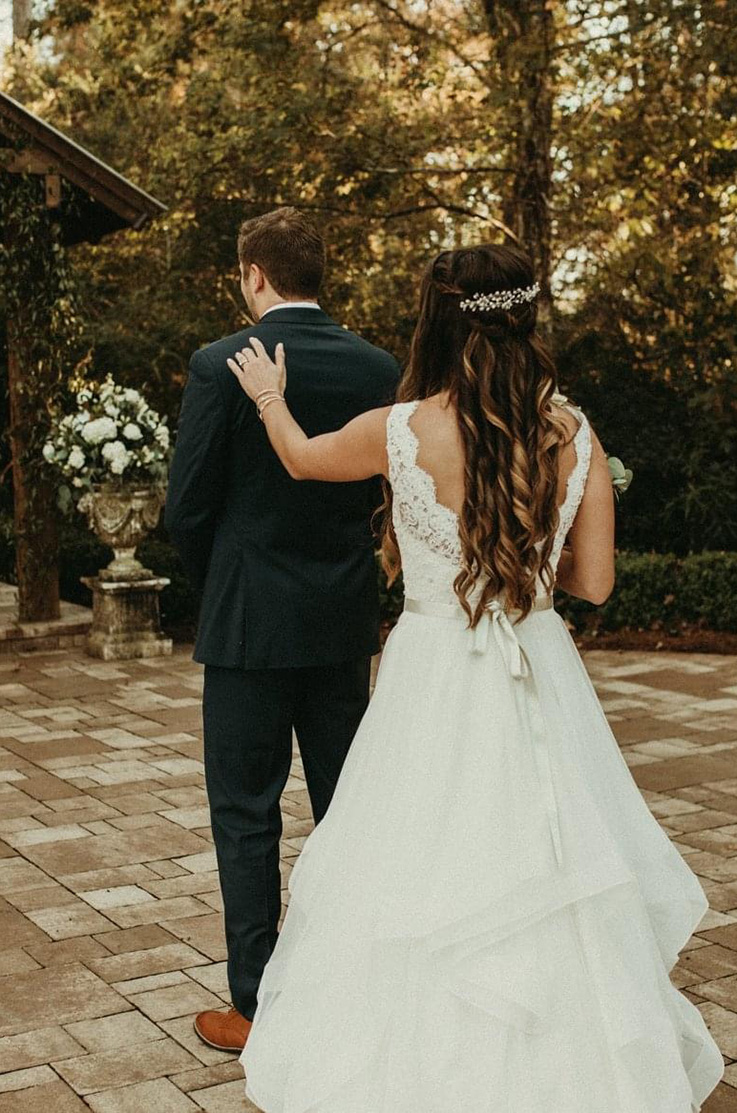 A bride in a tulle wedding dress taps the shoulder of a groom in a suit while both face away, while standing on a stone patio outdoors.