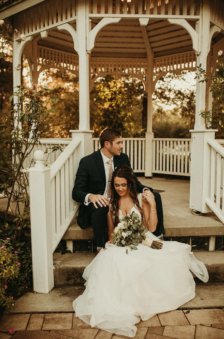 A bride in a tulle wedding dress and a groom in a suit sit on the steps of an outdoor gazebo, the bride has a bouquet in her lap.
