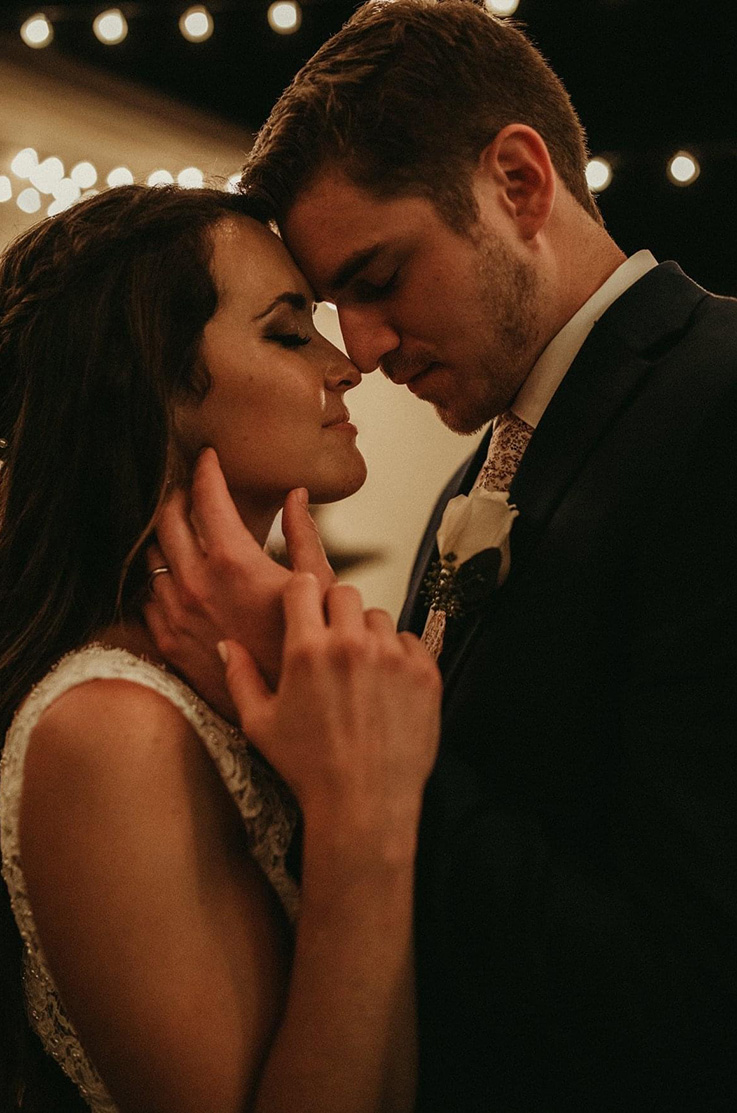 A close up of a bride in a wedding dress and a groom in a suit lean in close for a kiss, while standing under string lights at night.