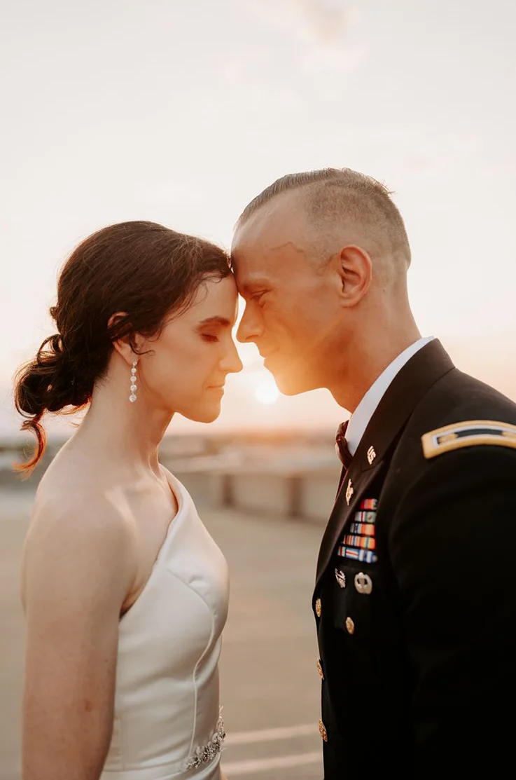 Bride in a one-shoulder fitted gown with a crystal belt, facing her groom as the sun sets on a rooftop.