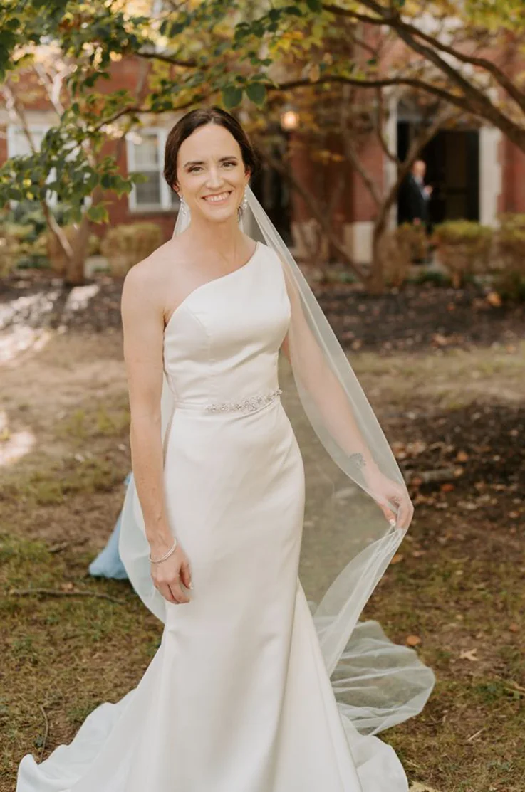 Smiling bride in a one-shoulder satin wedding dress with fit and flare skirt and a veil, standing by a tree.