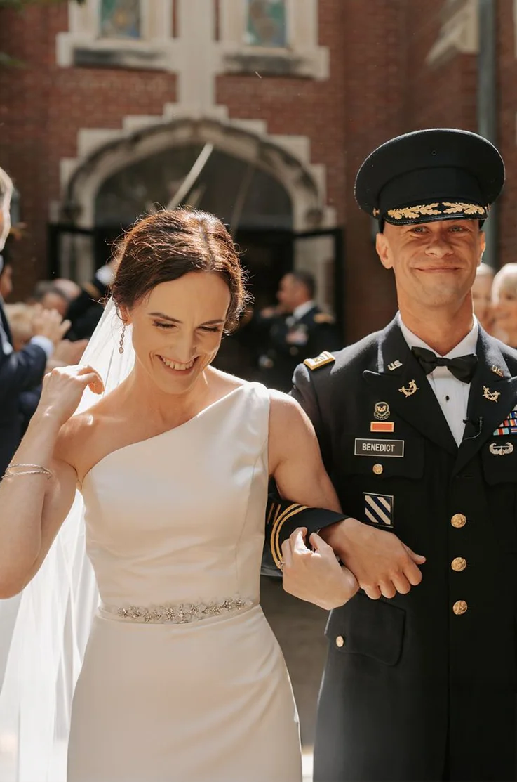 Close-up of bride in a one-shoulder satin gown with beaded belt and a veil, walks beside her groom in uniform, linking arms.