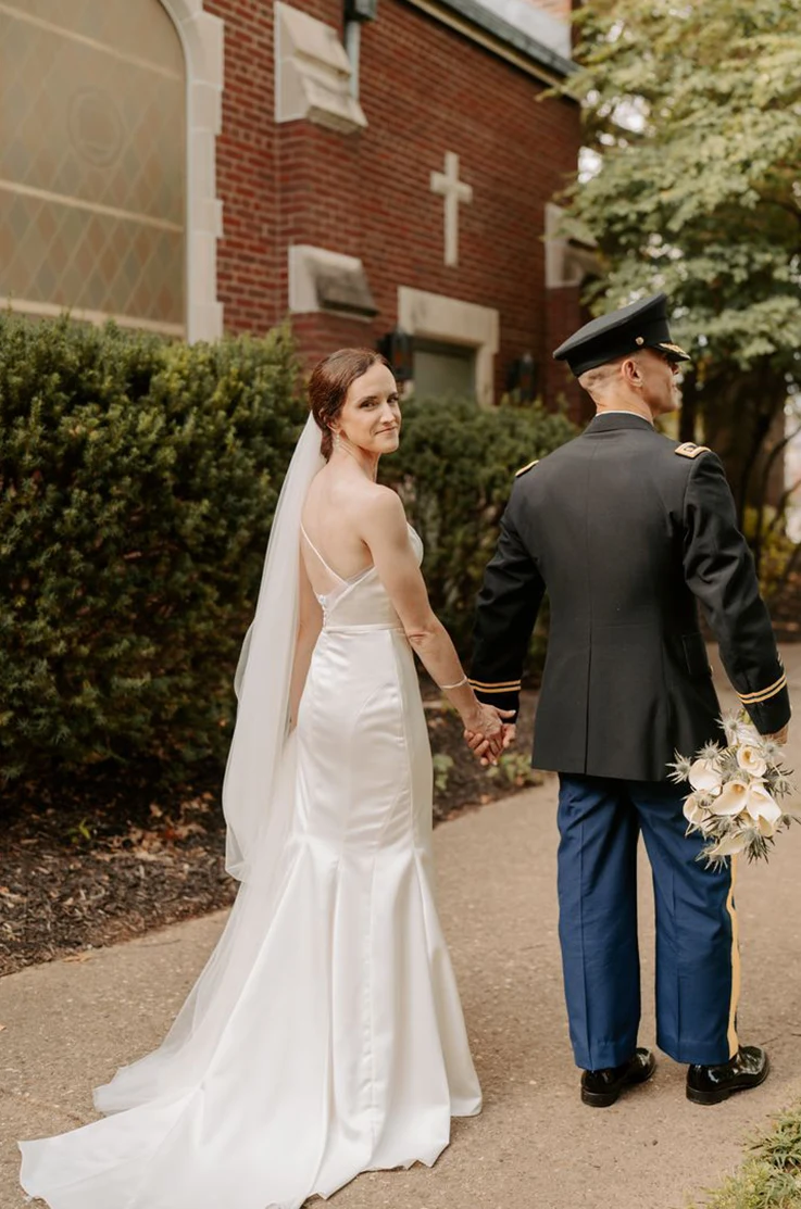 Bride looks over her shoulder in a satin fit and flare gown with a veil, as she holds hands with groom holding a bouquet.