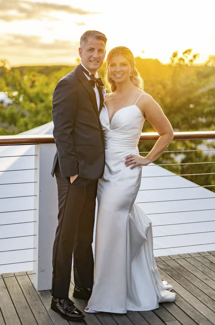 Bride in a draped bodice satin gown with fit and flare skirt and a sweetheart neckline, poses beside groom on a terrace at sunset.
