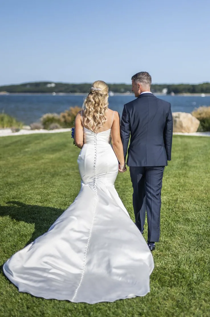 Back of bride in a buttoned satin gown with fit and flare skirt and thin straps, walking hand in hand with her groom at a waterfront.