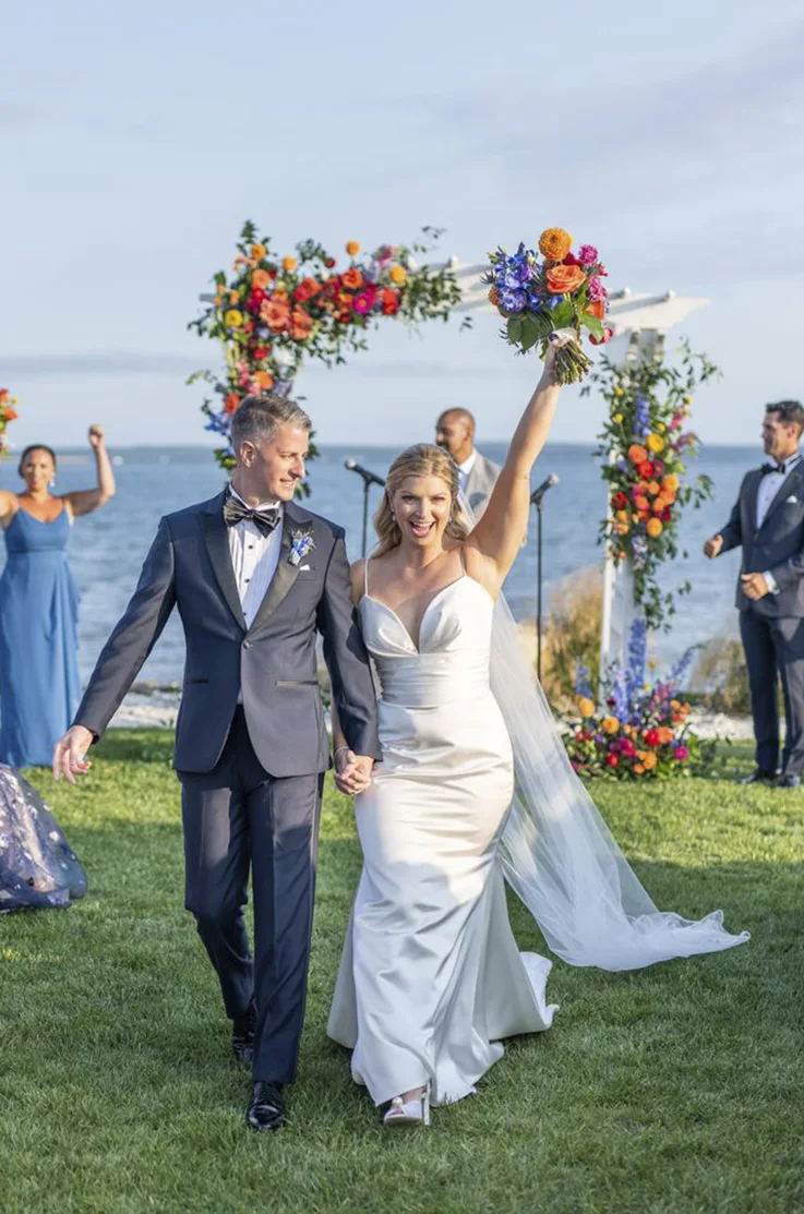 Bride in a draped satin spaghetti strap gown, holdng up a bouquet as she walks down the aisle with groom under a floral arch.