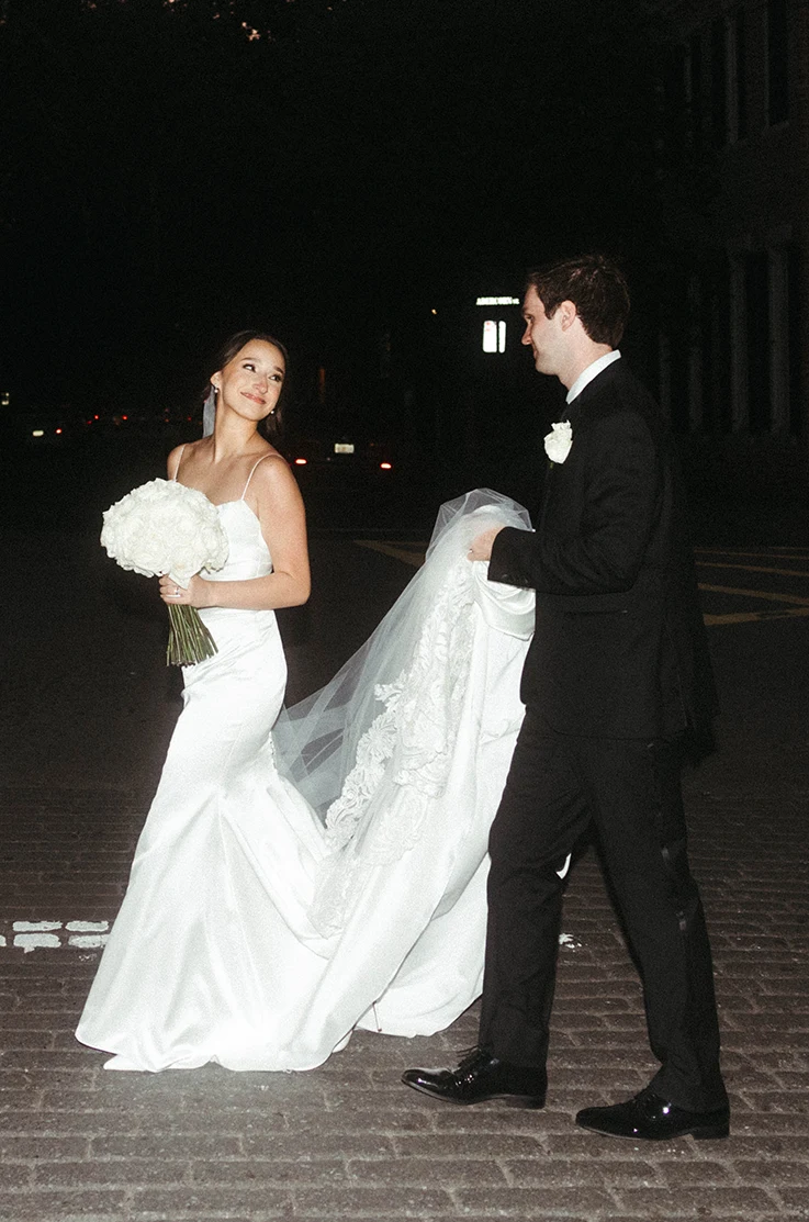 Groom holds up his bride's train in a satin fit and flare wedding dress, holding a white bouquet on a city street at night.