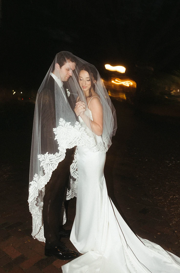 Bride in a satin fit and flare gown with thin straps and a lace-edge veil, embracing groom, in intimate moment at night.