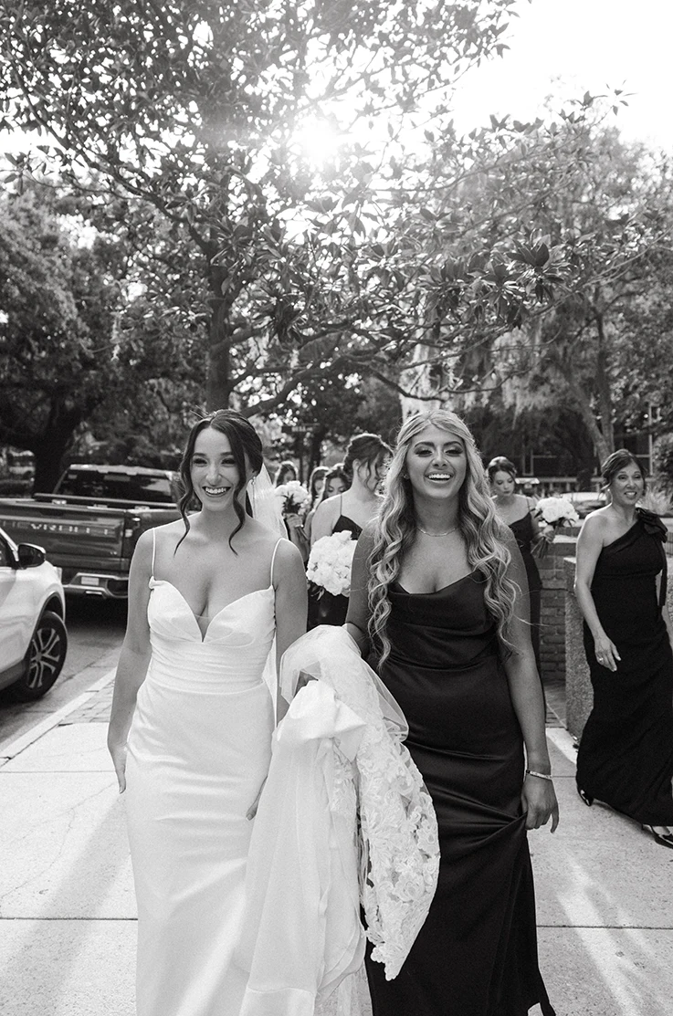 Bride in a plunging neck satin gown with a lace-trim veil, walks with her bridesmaids on city street, in black and white.