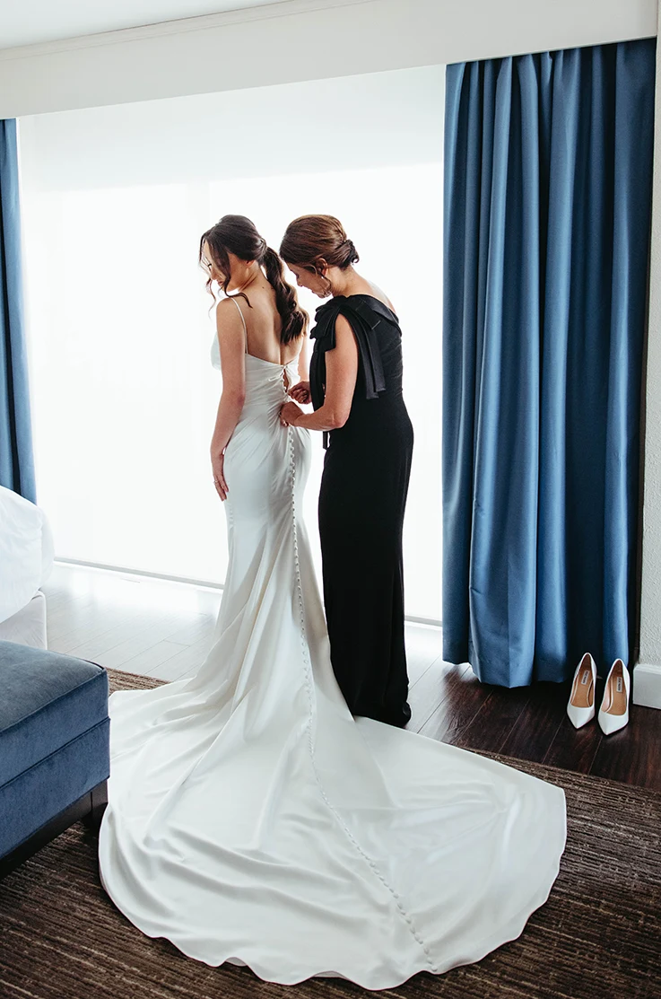 ack of bride being buttoned into a satin fit and flare gown with a low back, in a bridal suite by one of her bridesmaids.