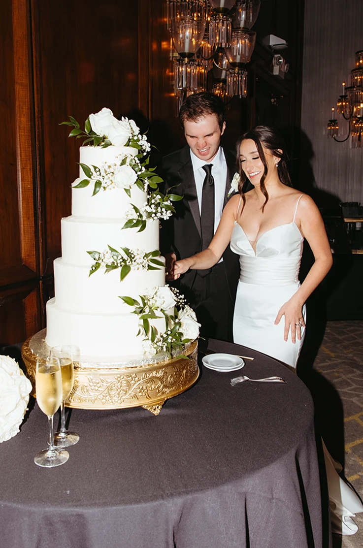 Bride in a satin draped gown with fit and flare skirt, cutting cake with her groom at indoor reception.