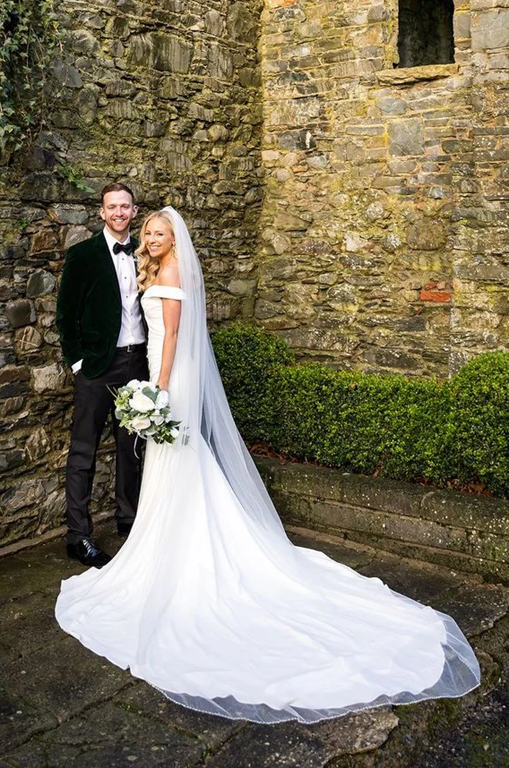 Bride in an off-the-shoulder crepe wedding dress with draping and a veil, posing beside groom in front of a stone building. 