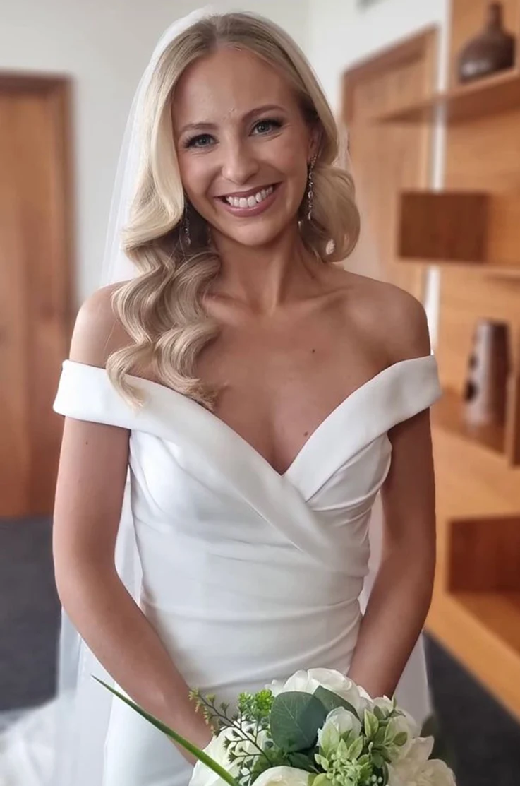 Close-up of smiling bride in an off-shoulder V-neck gown with a draped crepe bodice and a veil, is holding flowers indoors.