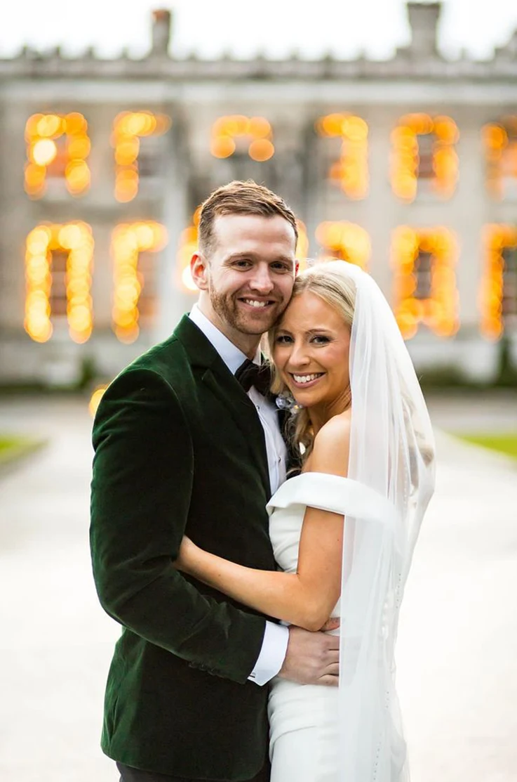 Groom holds bride in an embrace in an off-shoulder draped bodice gown in crepe with a veil, smiling in front of the venue.