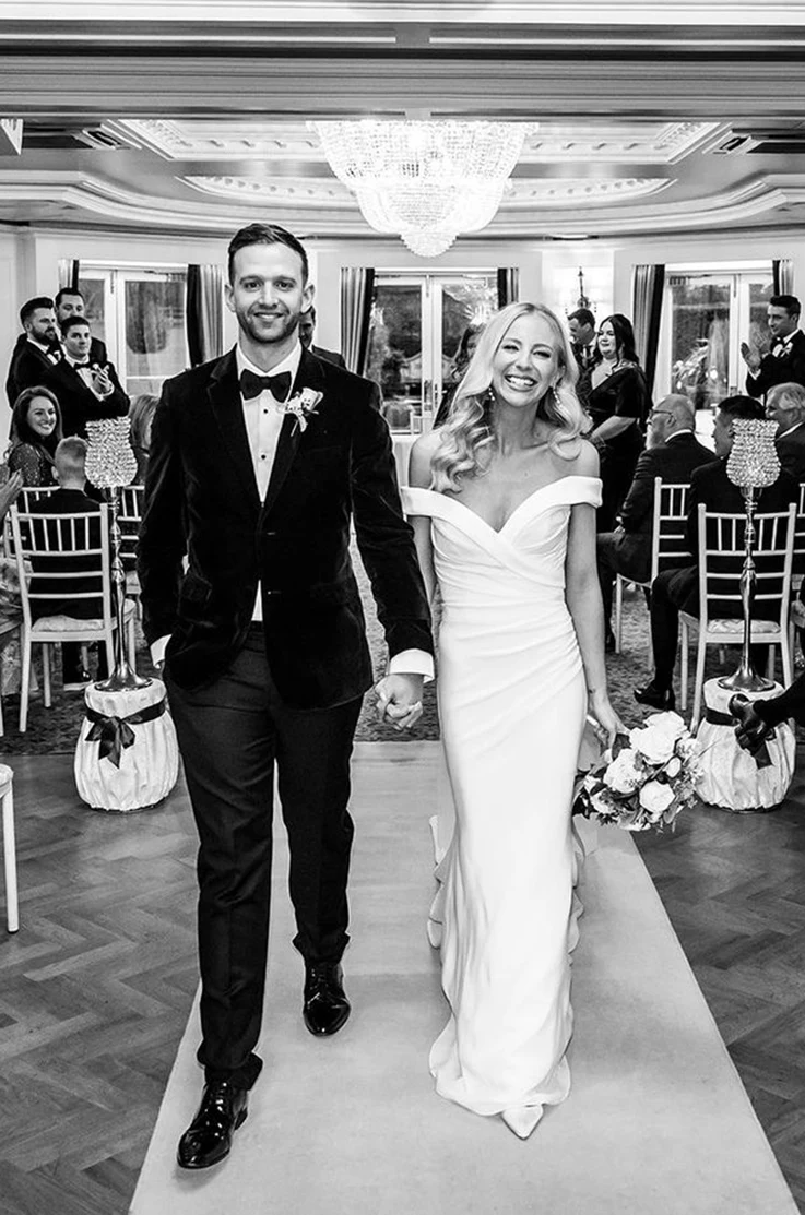 Bride in a draped crepe bodice gown with a off-shoulder neckline, walks hand in hand with groom in the aisle in black and white. 