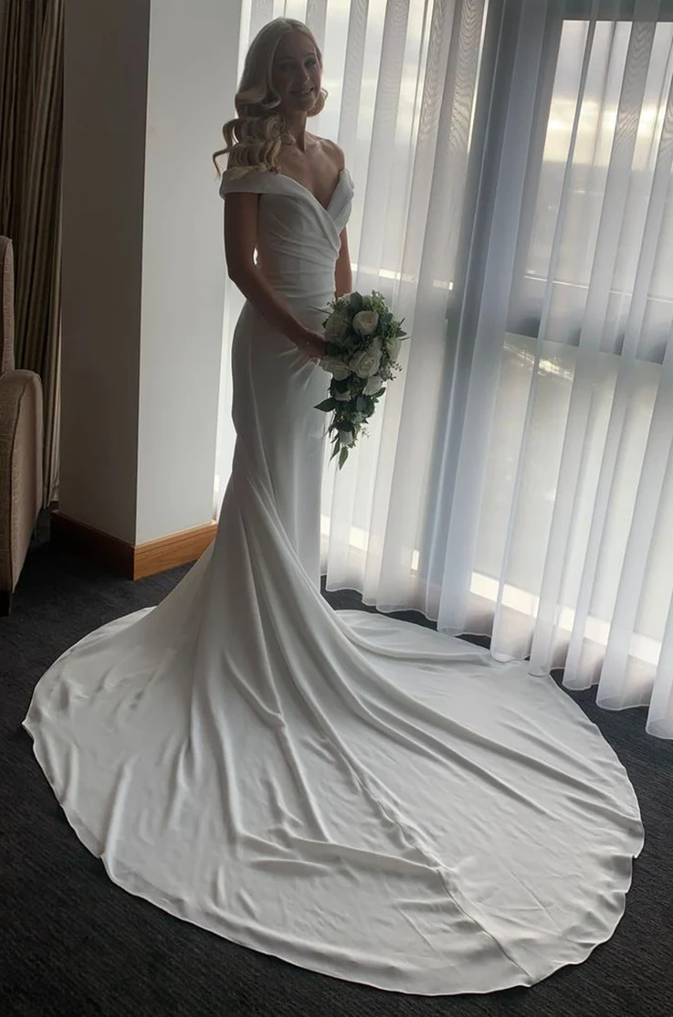 Bride in an off-shoulder draped gown in crepe, and is holding a bouquet by a bright window with white curtains.