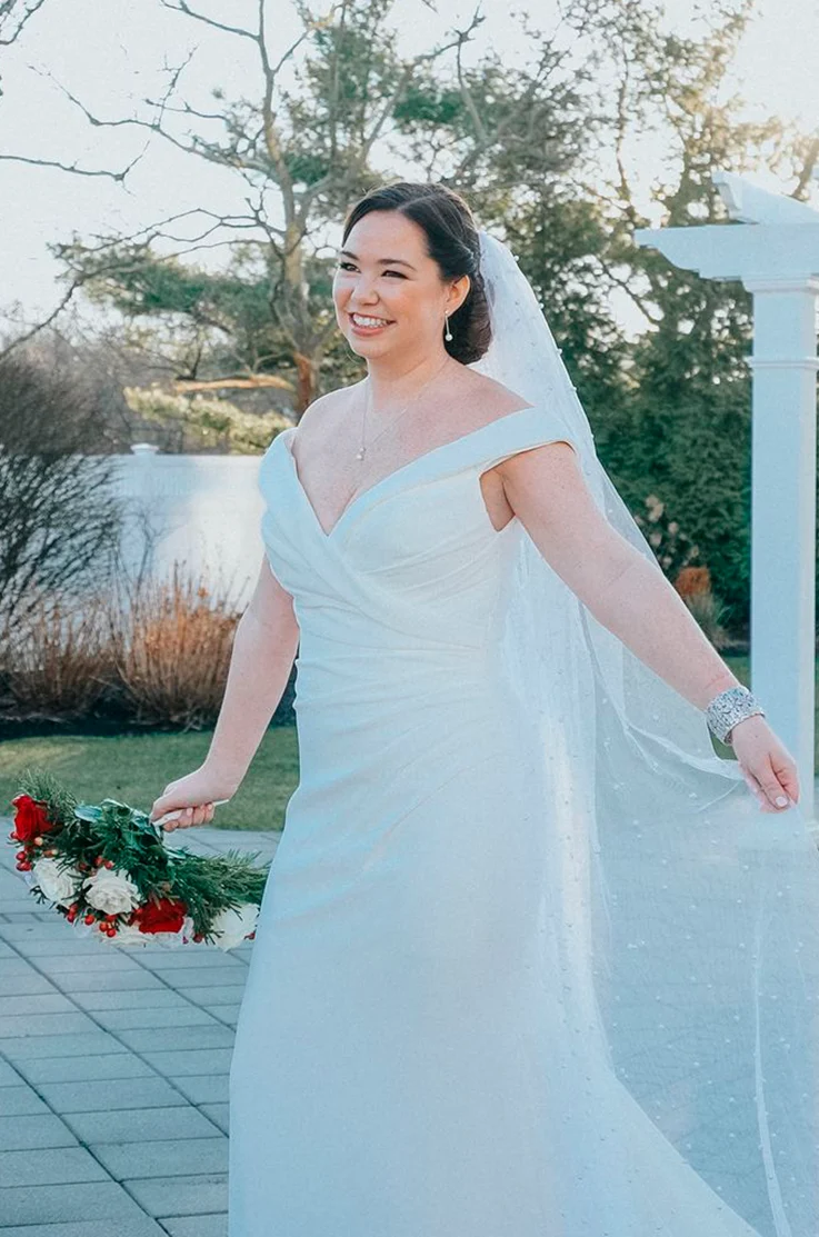 Bride in an off-shoulder fit and flare gown with a draped crepe bodice, V-neck and pearl veil, posing outdoors.